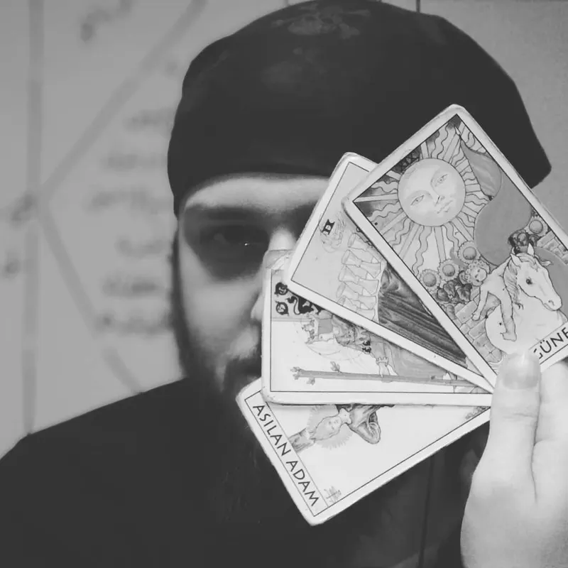 Oğuzhan Yavuz üç tarot kartını yelpaze gibi yüzünün önünde tutuyor, arkada astroloji çizimi