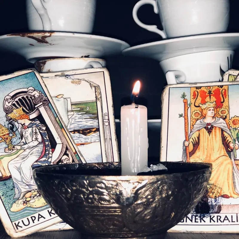 İki dik tarot kartının arasında gümüş kâsede beyaz mum, arkasında üst üste dizilmiş kahve fincanları